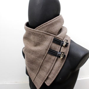 May include: A brown herringbone patterned scarf with black leather accents and silver-tone toggle closures. The scarf is displayed on a black mannequin, showcasing its design and texture. This accessory is designed to be worn around the neck.
