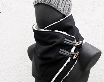 May include: A black cowl scarf with a white faux-shearling lining, featuring two black faux-leather tabs secured by brown buttons. The scarf is worn with a grey ribbed knit beanie cap on a black mannequin head and torso against a white wall.