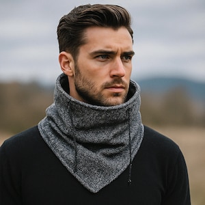 Pode incluir: Um aquecedor de pescoço texturizado cinza e preto com fecho de cordão. O aquecedor de pescoço é feito de um material espesso e quente, projetado para proteger o pescoço e a parte inferior do rosto do frio. A pessoa está usando um suéter preto.