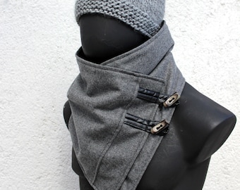May include: A grey knit beanie hat paired with a grey cowl neck scarf. The scarf features an asymmetrical wrap design with two black faux leather straps secured by light-colored toggle buttons, creating a layered look around the neck area.