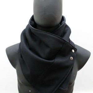 May include: A solid black cowl scarf featuring a structured, layered design that wraps around the neck. The scarf is secured on the side with two polished silver-toned metal snap buttons, creating a clean, angular silhouette with visible stitching.