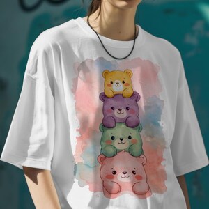 May include: White t-shirt featuring a stack of four cartoon teddy bears in yellow, purple, green, and pink. The bears have cute facial expressions and are stacked on top of each other. The background has a watercolor effect with pink, blue, and purple hues.
