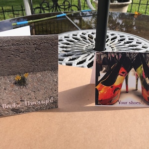 May include: Two greeting cards on a table. One card shows a flower growing through concrete with the text "Broke Through!". The other card displays a pair of high heels with the text "Your shoes..."