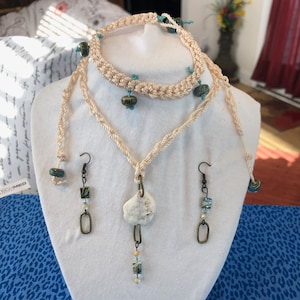 May include: A cream-colored macrame jewelry set displayed on a white mannequin. The set includes a necklace with a shell pendant, a bracelet, and earrings. The jewelry is adorned with blue and green beads and antique bronze-colored metal accents.