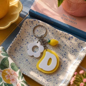 Puede incluir: Un llavero decorativo con una flor blanca, un limón amarillo y un dije de pera amarilla y blanca. El llavero está sobre un plato moteado azul y blanco. Un libro con el texto "Cote D'Azur" está en el fondo.