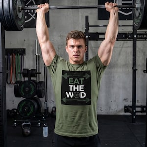 Könnte beinhalten: Ein olivgrünes T-Shirt mit dem Text "EAT THE WOD" in Weiß und Grün, getragen von einer Person, die eine Langhantel mit Gewichten in einem Fitnessstudio hebt. Die Person trägt schwarze Shorts.