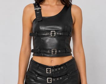 May include: A black faux leather crop top featuring an asymmetrical shoulder strap and two horizontal buckled straps across the chest, paired with matching black faux leather shorts that include two integrated buckled belts at the waistline.