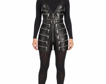 May include: A black leather romper featuring a deep V-neckline, thick shoulder straps with metal buckles, and a central front zipper. The garment is adorned with multiple horizontal silver-toned zippers across the bodice and shorts, and a waist belt.