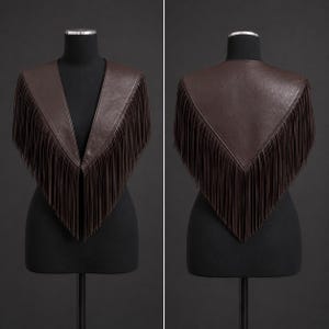 May include: A dark brown leather shawl featuring a V-shaped neckline and long, flowing fringe trim along the edges. The garment is displayed on a mannequin, showing both the front view with open panels and the back view with a pointed hemline.