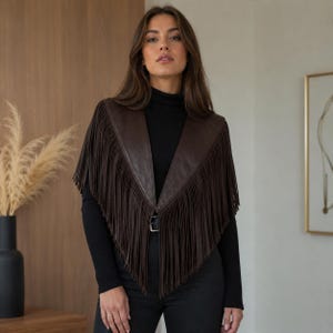 May include: A dark brown leather capelet featuring long, flowing fringe detailing along the edges and a single silver-toned buckle closure at the front. The garment is worn over a black long-sleeved ribbed turtleneck top and black trousers.