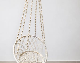 May include: A cream-colored macrame hanging chair featuring a circular frame with a woven net seat and backrest. The chair is suspended by four thick, twisted rope cords that meet at the top, and the bottom edge is finished with long fringe tassels.