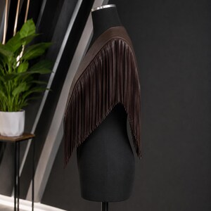 May include: A dark brown shoulder wrap featuring long, vertical fringe detailing that extends from the neckline down to the mid-torso. The garment is displayed on a black mannequin, showcasing a smooth, solid-colored texture and a clean, draped silhouette.