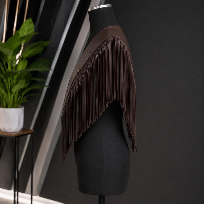 May include: A dark brown shoulder wrap featuring long, vertical fringe detailing that extends from the neckline down to the mid-torso. The garment is displayed on a black mannequin, showcasing a smooth, solid-colored texture and a clean, draped silhouette.