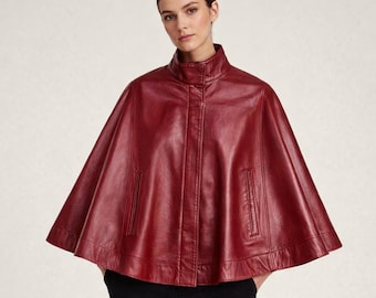 May include: A deep red leather cape featuring a high stand-up collar, a front button placket, and two vertical slit pockets. The garment has a wide, flared silhouette that drapes from the shoulders, providing a structured yet flowing appearance.