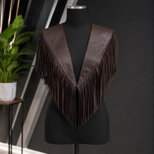 May include: A dark brown leather capelet featuring a deep V-neckline and long, thin fringe detailing that extends from the shoulders down to a pointed center. The garment is displayed on a black mannequin, showcasing its smooth texture and drape.