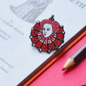 Può includere: Uno spillo in smalto rosso con un ritratto della regina Elisabetta I con una ruota zodiacale rossa e nera intorno al collo. Lo spillo è su un libro bianco con il testo "of the First ELIZABETH" visibile.