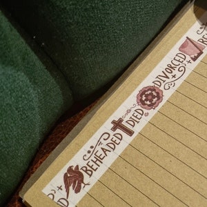 May include: A roll of decorative washi tape with a brown and white design. The tape features the words "Beheaded", "Died", "Divorced", "Survived", and "Beheaded" along with a cross and other decorative elements. The tape is partially unrolled and is lying on a brown notebook with a pen.