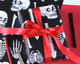 May include: A black gift box wrapped in skeleton and bone wrapping paper with a red satin ribbon. The wrapping paper features a repeating pattern of skeletons, bones, and anatomical illustrations.