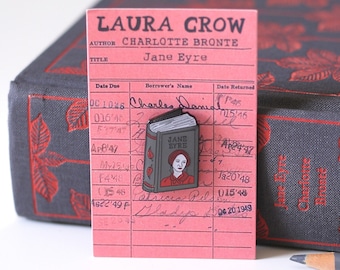 May include: A black and white enamel pin of a book titled "Jane Eyre" by Charlotte Bronte. The pin is on a pink library card with the text "Laura Crow" and "Charles Danial" written on it.