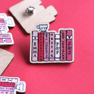 Peut inclure: Une épingle en bois représentant une pile de livres avec des titres comme "Emma", "Persuasion", "Mansfield Park", "Austen", "P&P", "Northanger Abbey" et "S&S". Une petite tasse à thé est posée sur le dessus des livres.