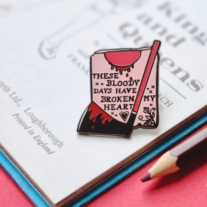 May include: A pink and black enamel pin with a red dripping heart and the text "These Bloody Days Have Broken My Heart".