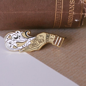 May include: A gold-colored enamel pin in the shape of a hand holding a bottle. The pin features the text "Lord, What Fools These Mortals Be" and a decorative swirl design in white and tan.