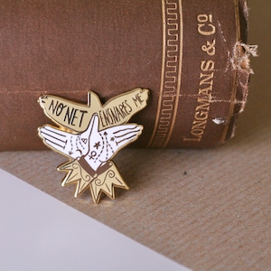 May include: Gold-tone enamel pin featuring a stylised bird with outstretched wings. The bird's wings are formed by two hands with the text "NO NET ENSNARES ME" written on the wings.
