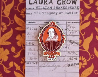 May include: A pin featuring a portrait of William Shakespeare in an oval frame with a red and orange border. The pin is placed on a library card with the title "The Tragedy of Hamlet" and the author's name, William Shakespeare.