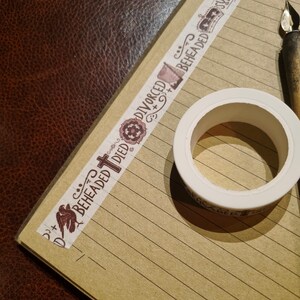 May include: A close-up of a brown and white washi tape with the words "Beheaded Died Divorced Beheaded" printed on it. The tape is on a lined notebook page with a white roll of tape and a pen.
