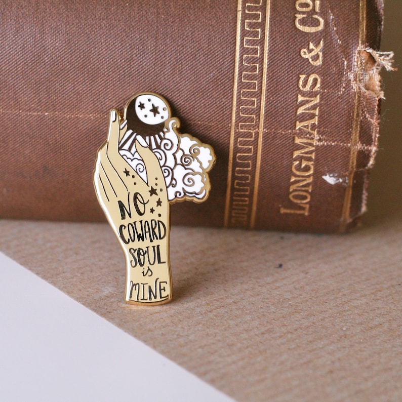 May include: Gold-toned enamel pin featuring a hand holding a crescent moon and stars with the text "No Coward Soul is Mine".