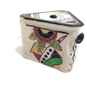 May include: A white ceramic teapot with a triangular shape and a colorful geometric design. The teapot has a spout and a lid with a blue knob. The design features a red, yellow, green, and blue floral pattern.