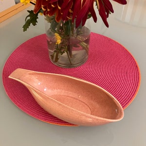 May include: A pink ceramic serving dish in the shape of a boat, sitting on a pink and orange circular placemat.