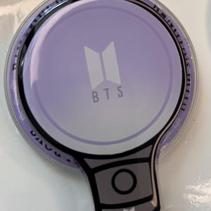 May include: A light purple BTS light stick with a white logo and the letters "BTS". The handle is gray with a black circle and a small white logo. The light stick is on a white surface.