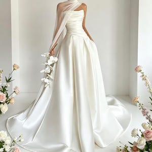Peut inclure: Une robe de mariée en satin blanc avec un corsage bustier et une jupe ample. La robe présente un détail d'épaule drapée et une longue traîne. La mariée tient un bouquet d'orchidées blanches. Thème : mode nuptiale.