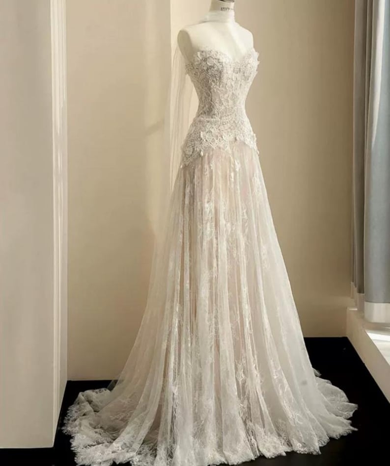 Peut inclure: Robe de mari&eacute;e blanche sur mannequin. La robe pr&eacute;sente un d&eacute;collet&eacute; en c&oelig;ur et est orn&eacute;e de dentelle complexe sur le corsage et la jupe. La jupe descend jusqu'au sol, cr&eacute;ant une silhouette romantique et &eacute;l&eacute;gante. Un voile transparent est drap&eacute; sur la t&ecirc;te du mannequin.