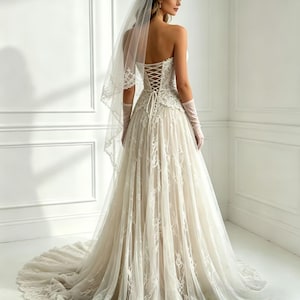 Peut inclure: Robe de mari&eacute;e ivoire sans bretelles avec dentelle d&eacute;taill&eacute;e et jupe fluide. Le dos pr&eacute;sente une fermeture de style corset. Un voile transparent et de longs gants compl&egrave;tent le look de mari&eacute;e. La robe a une longue tra&icirc;ne.