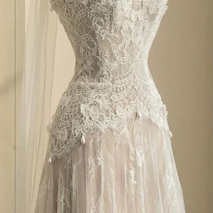 Peut inclure: Robe de mari&eacute;e en dentelle blanche sans bretelles sur mannequin. La robe pr&eacute;sente des d&eacute;tails de dentelle florale complexes, un corsage ajust&eacute; et une jupe fluide. De d&eacute;licats accents de perles ornent le corsage et la jupe, et un voile transparent drape le mannequin.