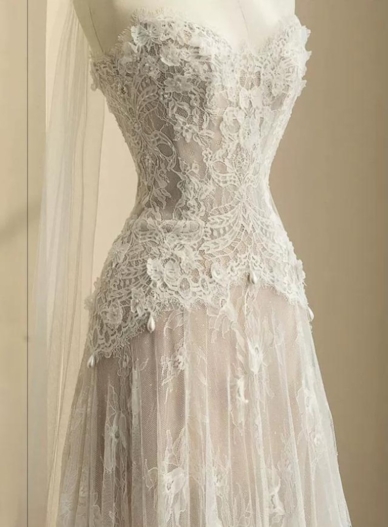 Peut inclure: Robe de mari&eacute;e en dentelle blanche sans bretelles sur mannequin. La robe pr&eacute;sente des d&eacute;tails de dentelle florale complexes, un corsage ajust&eacute; et une jupe fluide. De d&eacute;licats accents de perles ornent le corsage et la jupe, et un voile transparent drape le mannequin.