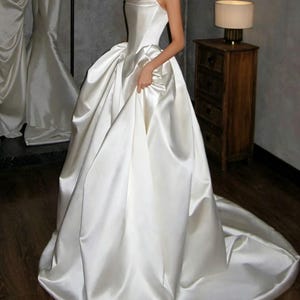 Peut inclure: Robe de mariée en satin blanc sans bretelles, avec une jupe ample et une longue traîne. La robe a un corsage ajusté et une jupe volumineuse qui descend jusqu'au sol. Le tissu a un éclat subtil.