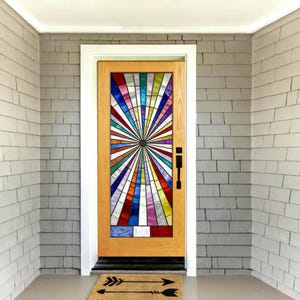 Op de afbeelding: Een houten deur met een glas-in-loodpaneel met een zonnestraalontwerp in verschillende kleuren, waaronder rood, blauw en geel. De deur heeft een zwarte handgreep en staat in een wit omlijste deuropening tegen een grijze bakstenen muur. Een welkomstmat met pijlen ligt op de grond.