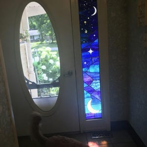 Puede incluir: Una puerta blanca con una ventana ovalada y un panel de vidrieras. El panel presenta un diseño de cielo nocturno con estrellas, una luna creciente y varios tonos de azul y morado. La puerta está en un pasillo bien iluminado.