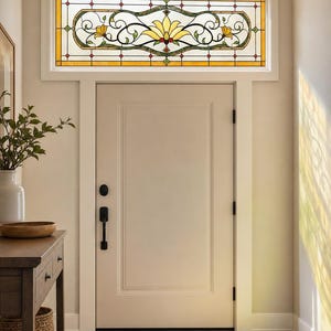 Könnte beinhalten: Ein rechteckiges Buntglas-Oberlicht mit einem zentralen gelben Blumenmotiv, grünen Ranken und roten Akzenten, eingefasst in einen weißen Rahmen über einer cremefarbenen Tür mit schwarzen Beschlägen und rechteckigem Paneel.