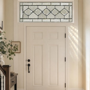 Può includere: Una porta d'ingresso bianca a sei pannelli sormontata da una finestra a specchiatura rettangolare. La specchiatura contiene vetri trasparenti testurizzati disposti in un motivo geometrico a rombi con una cornice in metallo scuro.