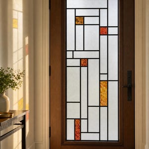 Könnte beinhalten: Eine dunkelbraune Holztür mit einem hohen, rechteckigen Glaseinsatz in geometrischem Gittermuster. Die Glasscheibe enthält mattweiße Abschnitte, die mit kleinen, strukturierten bernsteinfarbenen und orangefarbenen Glasquadraten durchsetzt sind.