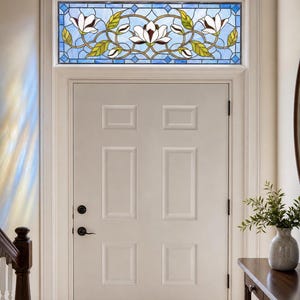Könnte beinhalten: Ein rechteckiges Buntglas-Oberlicht, das über einer weißen Tür mit sechs Paneelen installiert ist. Das Glas zeigt ein florales Design mit weißen Magnolienblüten, grünen Blättern und braunen Stielen vor einem Hintergrund aus hellblauen Glasscheiben.