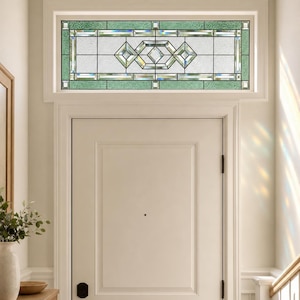 Op de afbeelding: Een rechthoekig glas-in-lood bovenlicht boven een witte deur. Het raam heeft een centraal geometrisch patroon met heldere geslepen glazen segmenten, omlijst door gestructureerde lichtgroene glazen randen en kleine vierkante accenten.