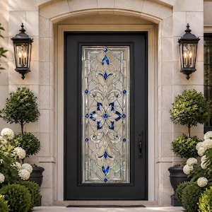 Op de afbeelding: Een zwarte voordeur met een decoratief glaspaneel met een bloemmotief met blauwe en heldere accenten. De deur wordt geflankeerd door twee zwarte lantaarns en potplanten, waardoor een elegante entree ontstaat.