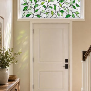 Könnte beinhalten: Ein rechteckiges Oberlicht aus Buntglas mit einem sich wiederholenden Muster aus grünen Blättern und Ranken vor einem klaren, strukturierten Glashintergrund. Das Design ist in einen weißen Rahmen über einer weißen Innentür mit schwarzen Beschlägen gefasst.