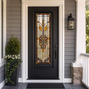 Puede incluir: Una puerta de entrada negra con una ventana decorativa de vidrieras con patrones geométricos en tonos naranja, amarillo y marrón. La puerta está enmarcada por molduras blancas y colocada contra un revestimiento gris. Una alfombra negra y una planta en maceta están frente a la puerta.