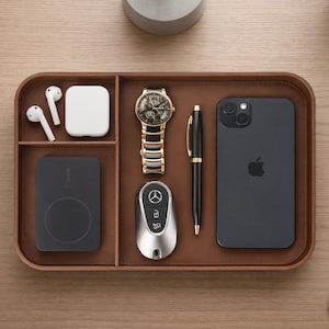 May include: A rectangular brown leather valet tray with rounded corners and three internal compartments. The tray contains a pair of white wireless earbuds, a white charging case, a black power bank, a gold and black watch, a car key fob, a pen, and a black smartphone.